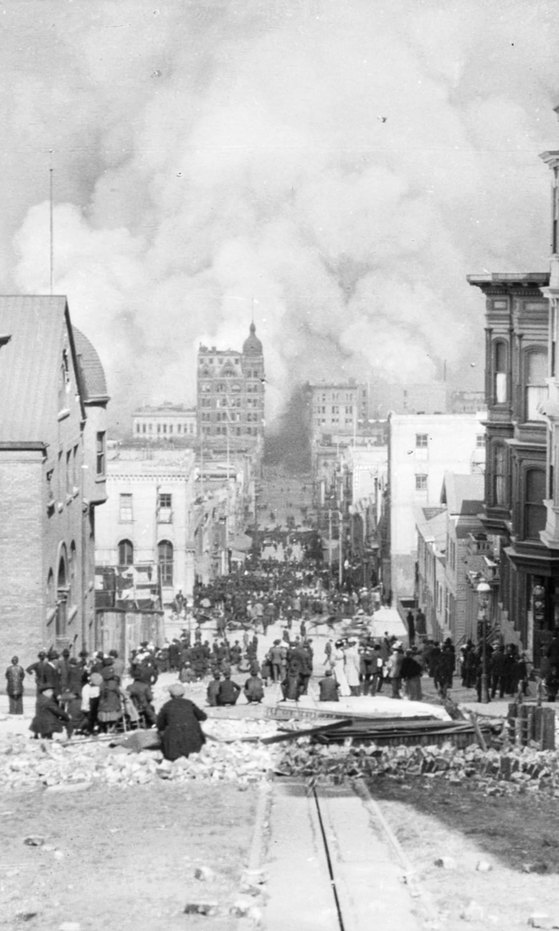 Sacramento Street on the morning of April 18, 1906: smoke from the fire towers over the city while people in the street, and on chairs by a ruined house, watch it come.